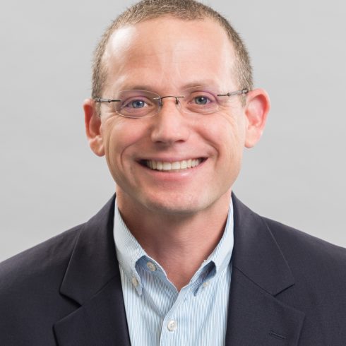 A person wearing glasses and a suit jacket smiles in a portrait against a gray background.