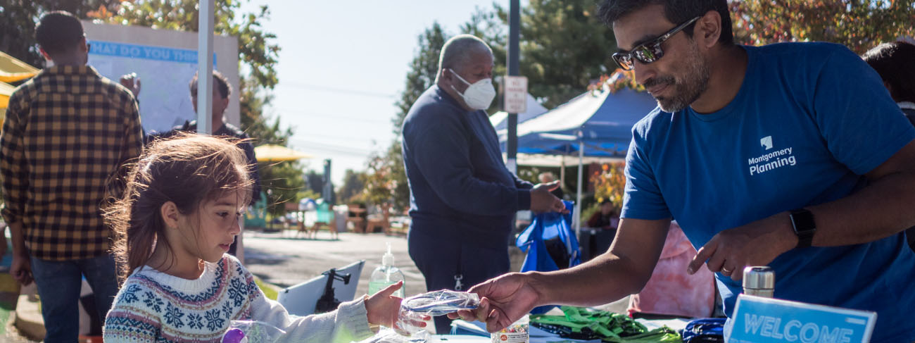 Montgomery Planning staff member hands giveaway sunglasses to a little girl at an outreach event