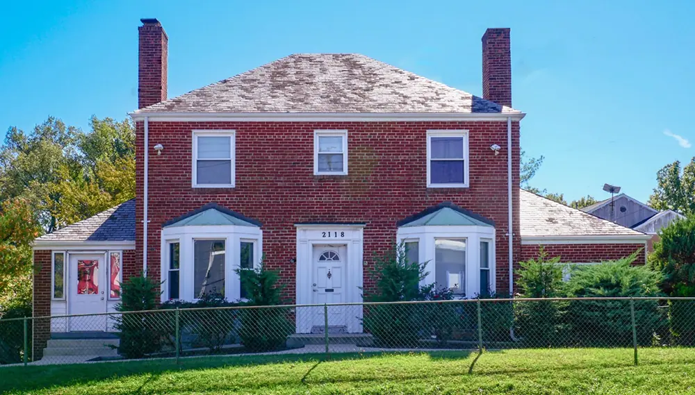 A charming two-story red brick house with two chimneys, a white door, and bay windows. Surrounded by green bushes and enclosed by a chain-link fence, it sits gracefully on a grassy lawn under the clear blue sky.
